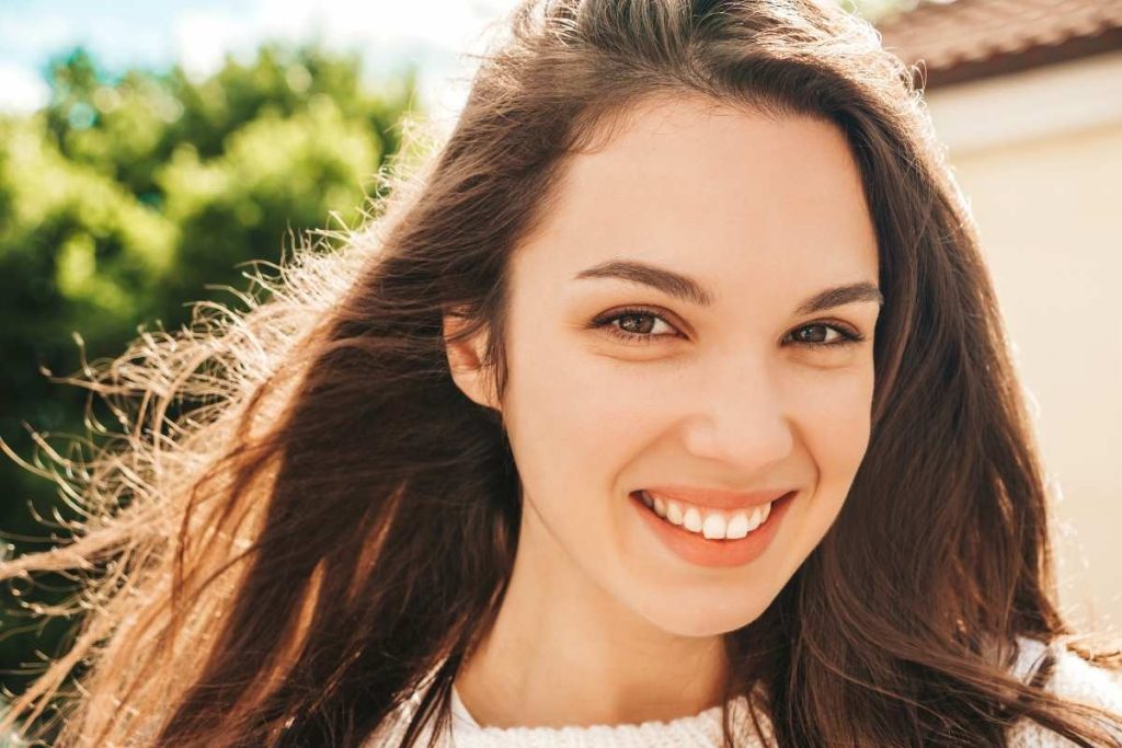 Smiling young woman with long brown hair outdoors in natural sunlight near greenery.