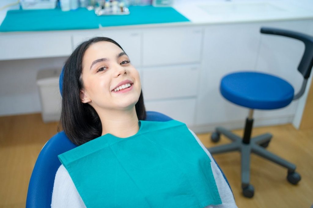 Smiling woman in dental bib at orthodontic office with dental chair visible in background.