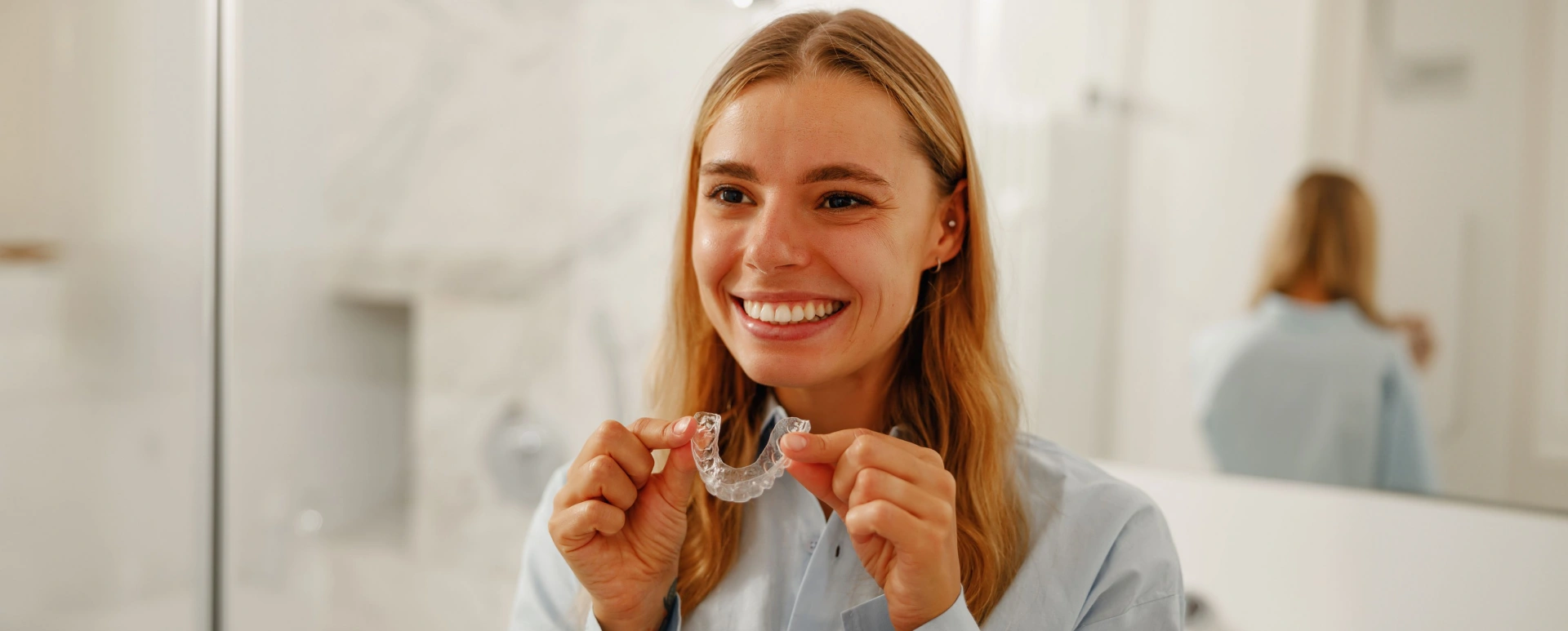 Woman smiling while holding clear orthodontic aligners in modern dental office setting.